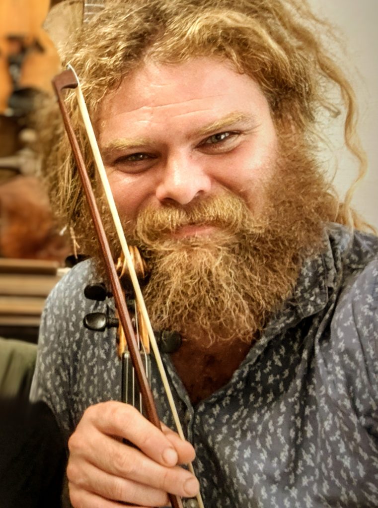 En skäggig man med lockigt hår ler medan han håller en fiol och en stråke nära sitt ansikte.