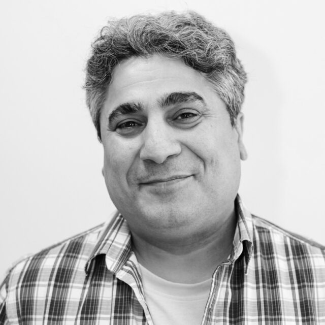 En man med grått hår ler, klädd i rutig skjorta, mot en vit bakgrund.