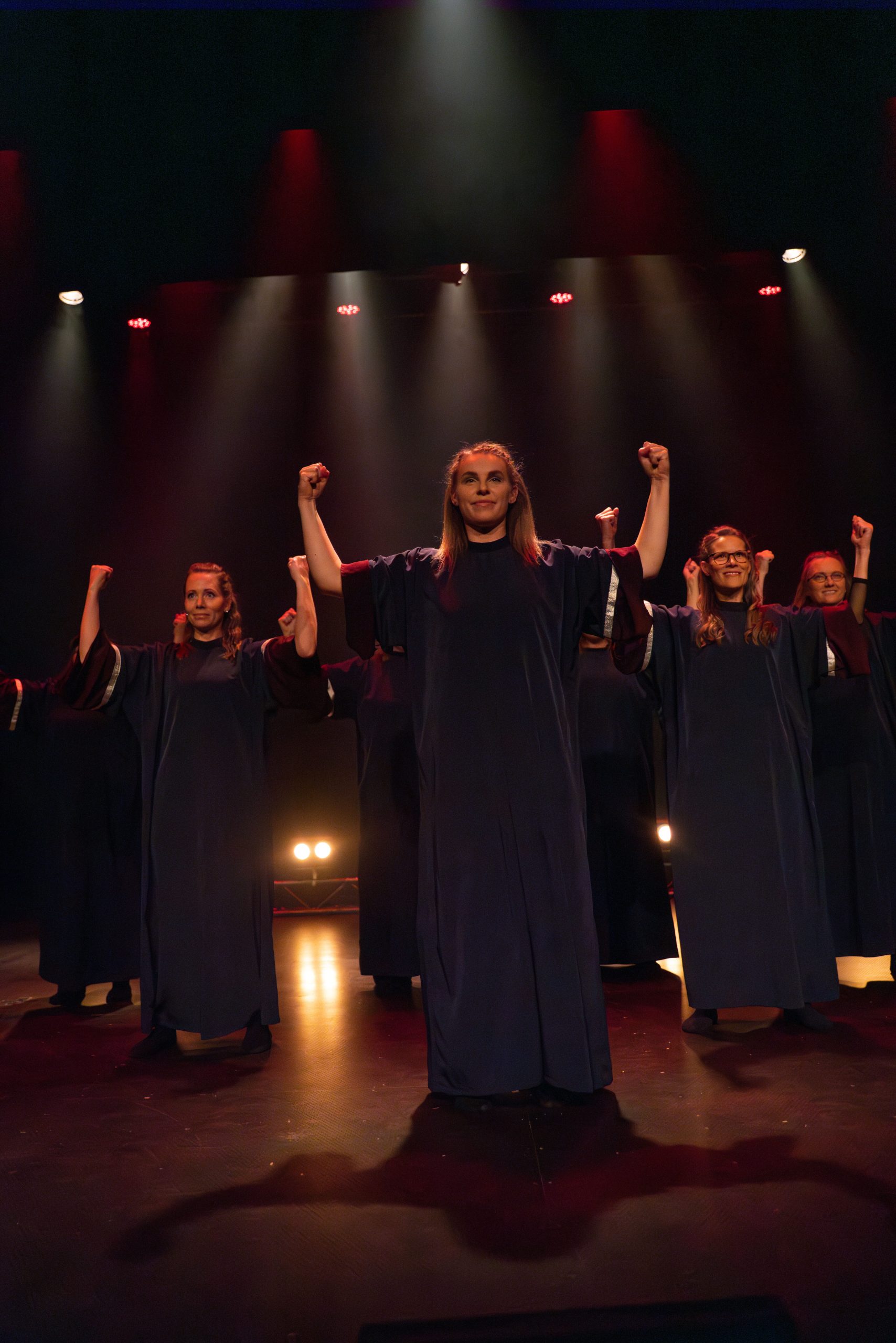En grupp mörkklädda personer står på scenen med höjda armar i en dramatisk ljussättning.