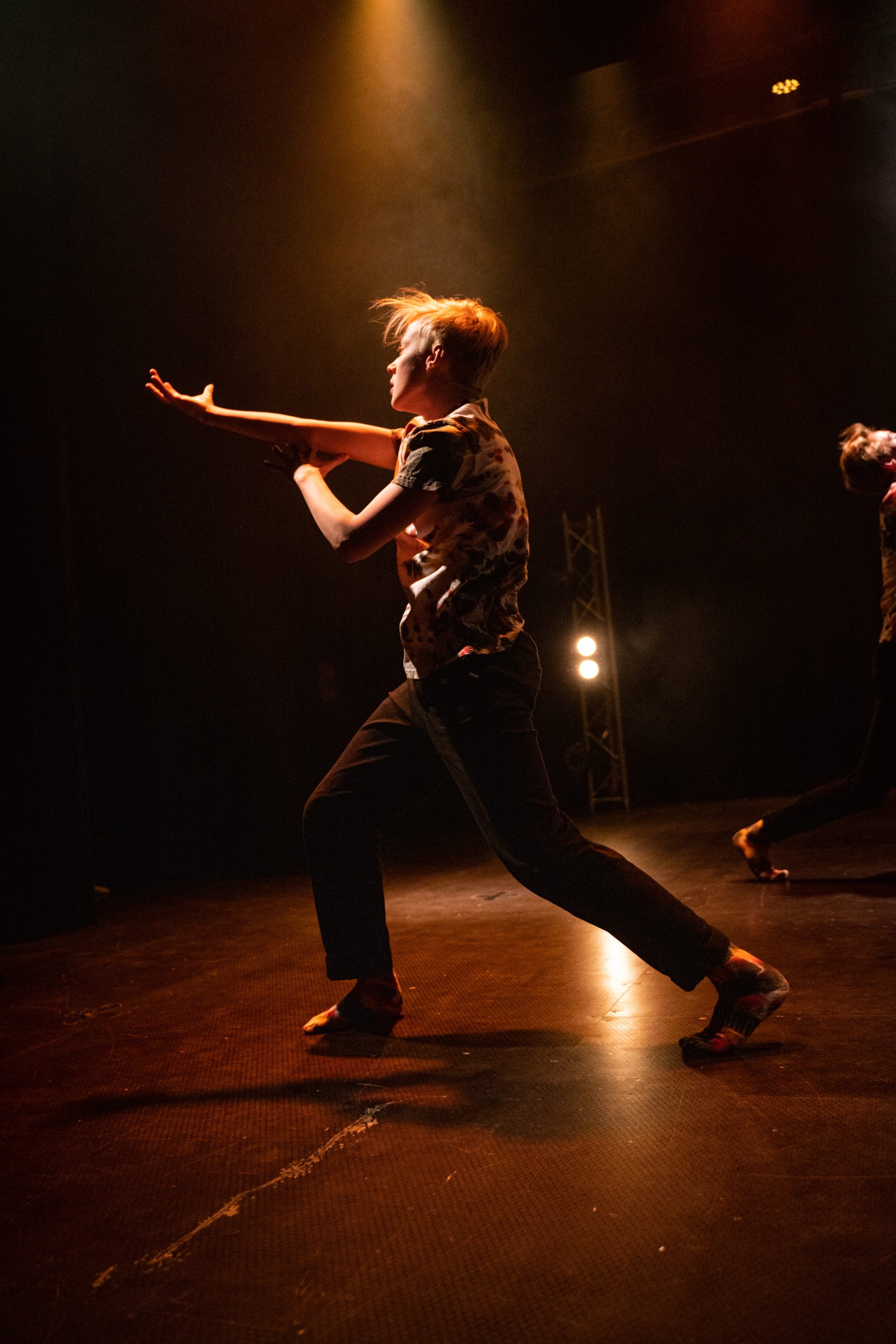 En person i mönstrad skjorta och mörka byxor dansar dramatiskt på scenen under varma strålkastare.