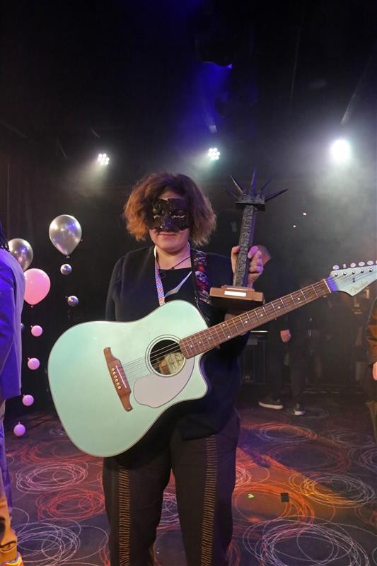 En maskerad person håller i en turkos gitarr och en spikförsedd trofé, med ballonger i bakgrunden.