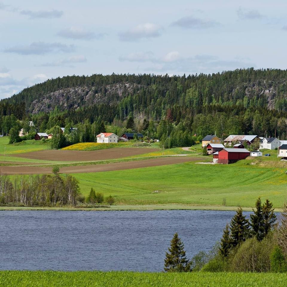 Ett lantligt landskap med gröna fält, utspridda hus, skogsklädda kullar och en sjö i förgrunden.