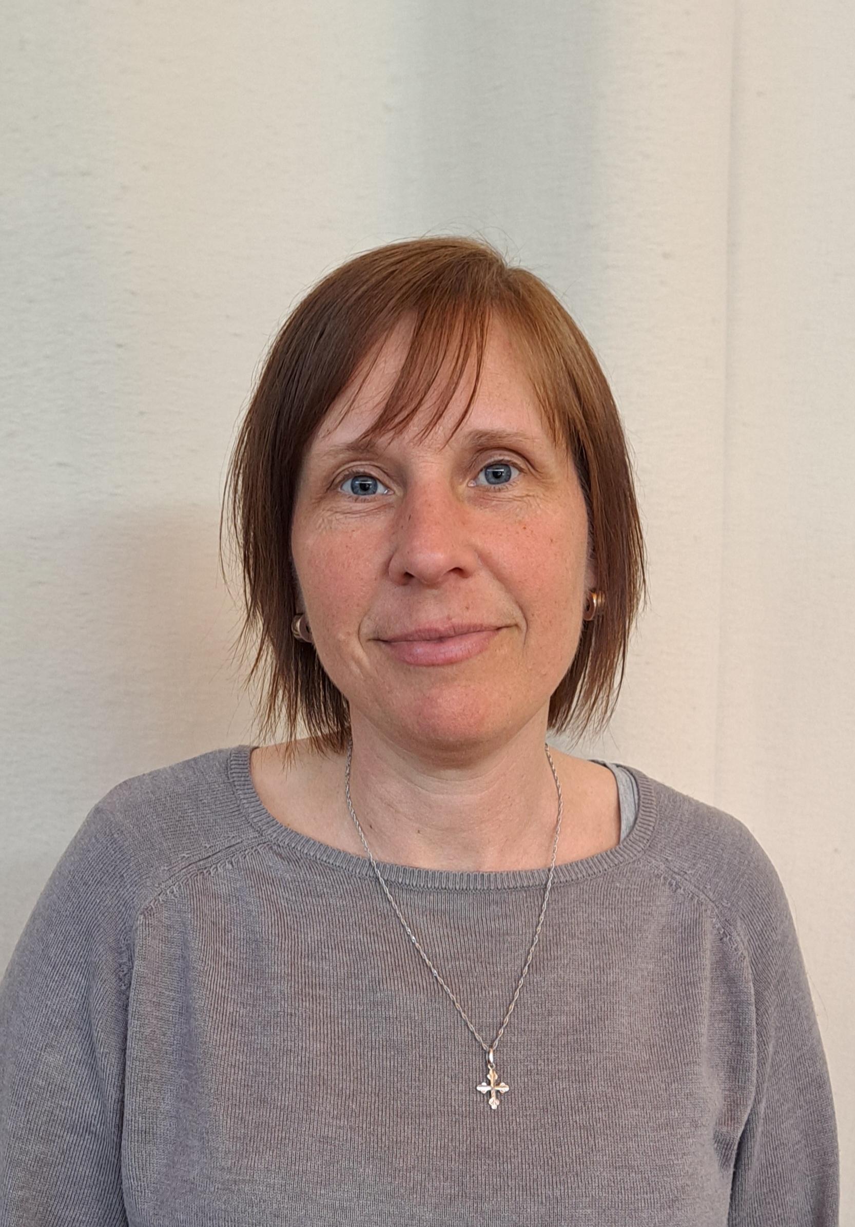 En kvinna med kort brunt hår, klädd i grå topp och korshalsband, står framför en ljus bakgrund.