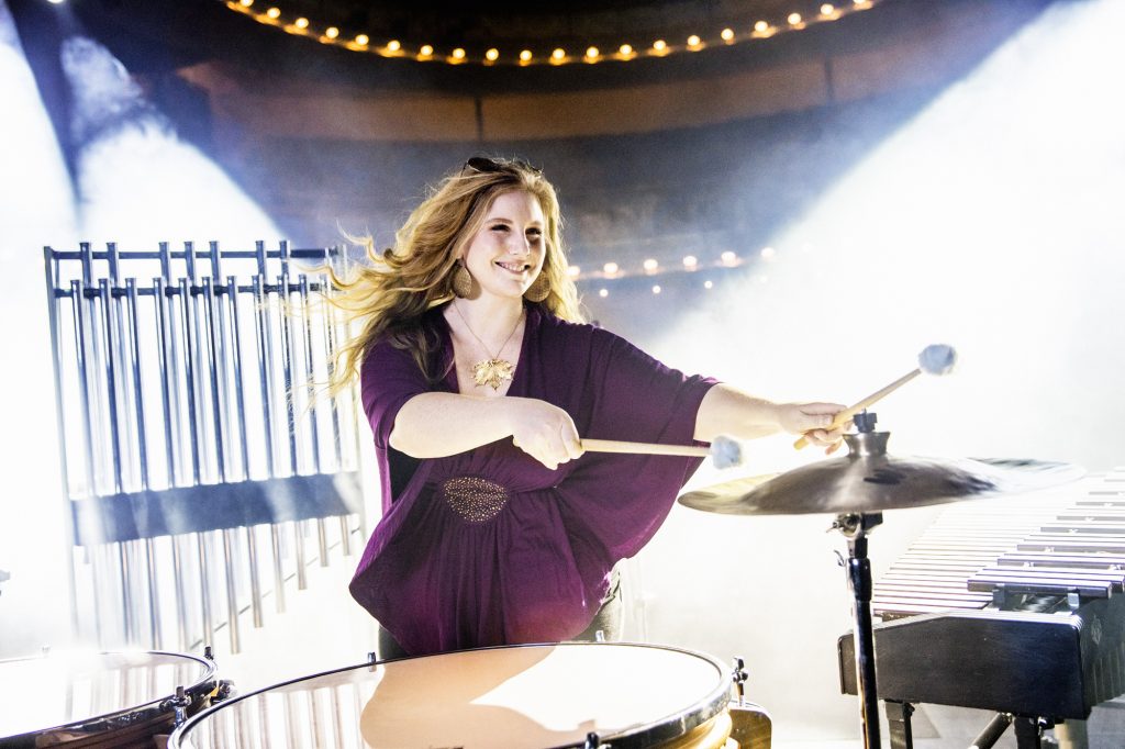 Kvinna i lila klänning spelar slagverksinstrument på scen med dramatisk belysning bakom sig.