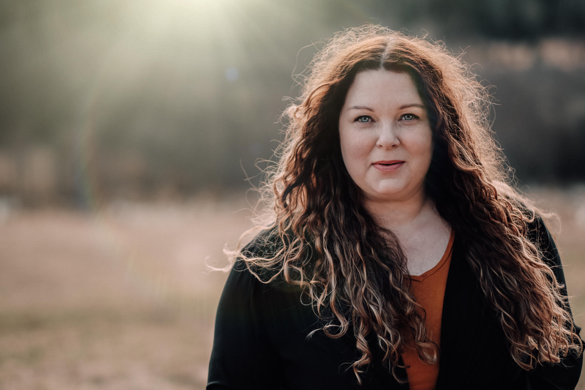 Kvinna med långt lockigt hår står utomhus i solljus, klädd i svart jacka och orange topp och tittar in i kameran.