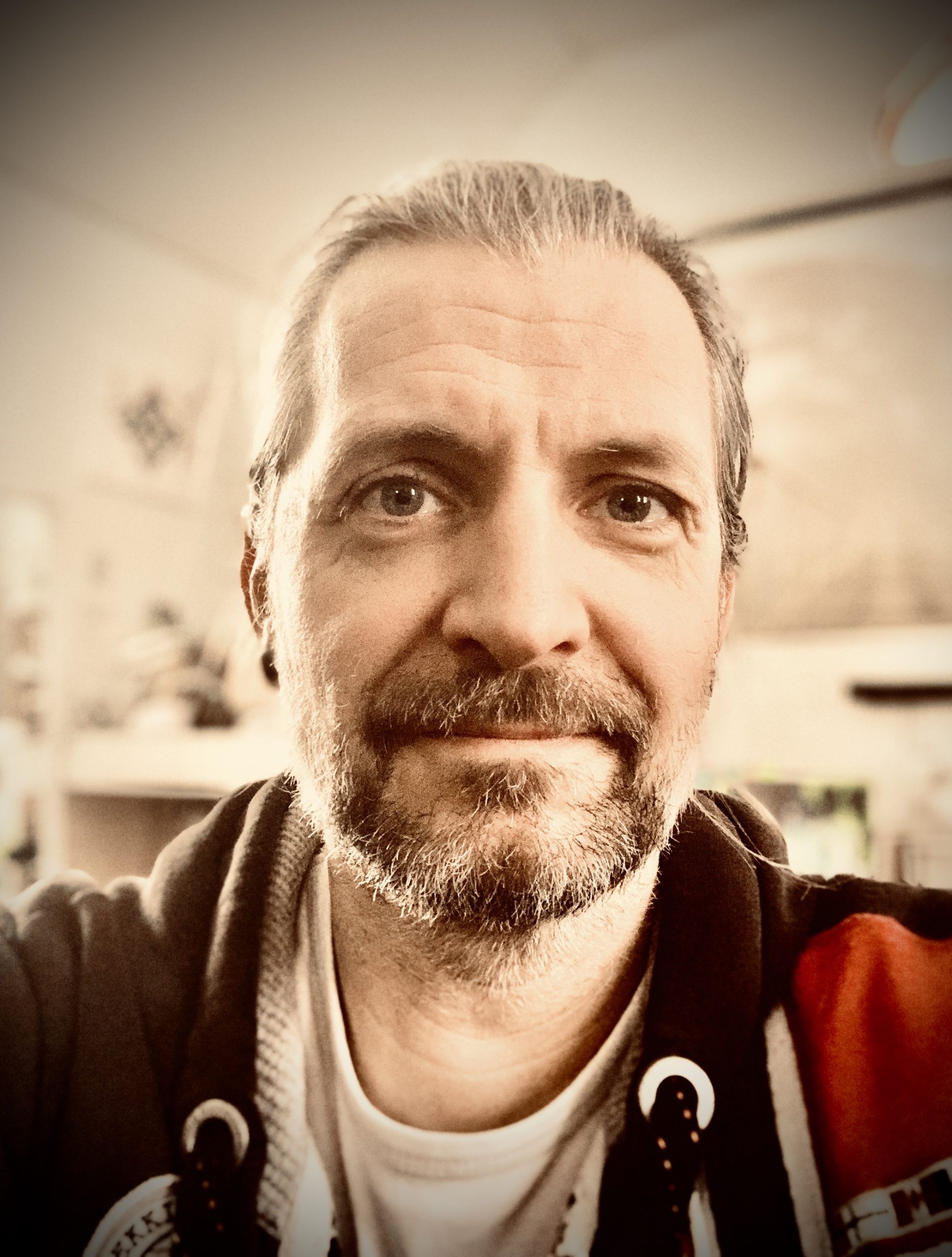 Leende man med skägg och mustasch, iklädd huvtröja, inomhus med suddig bakgrund.