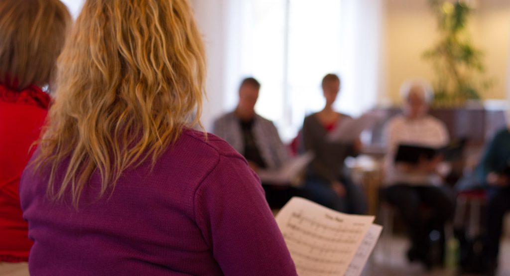 En person med noter i handen står framför en grupp människor som sitter i ett ljust rum och kanske repeterar.