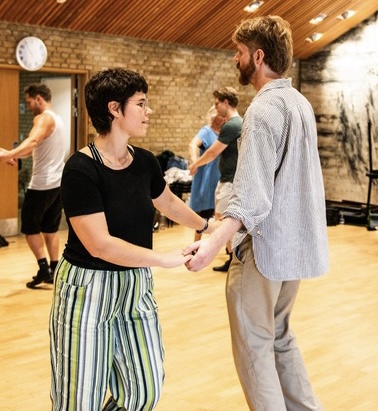 Två personer håller varandra i handen och dansar i en studio, med andra par som dansar i bakgrunden.