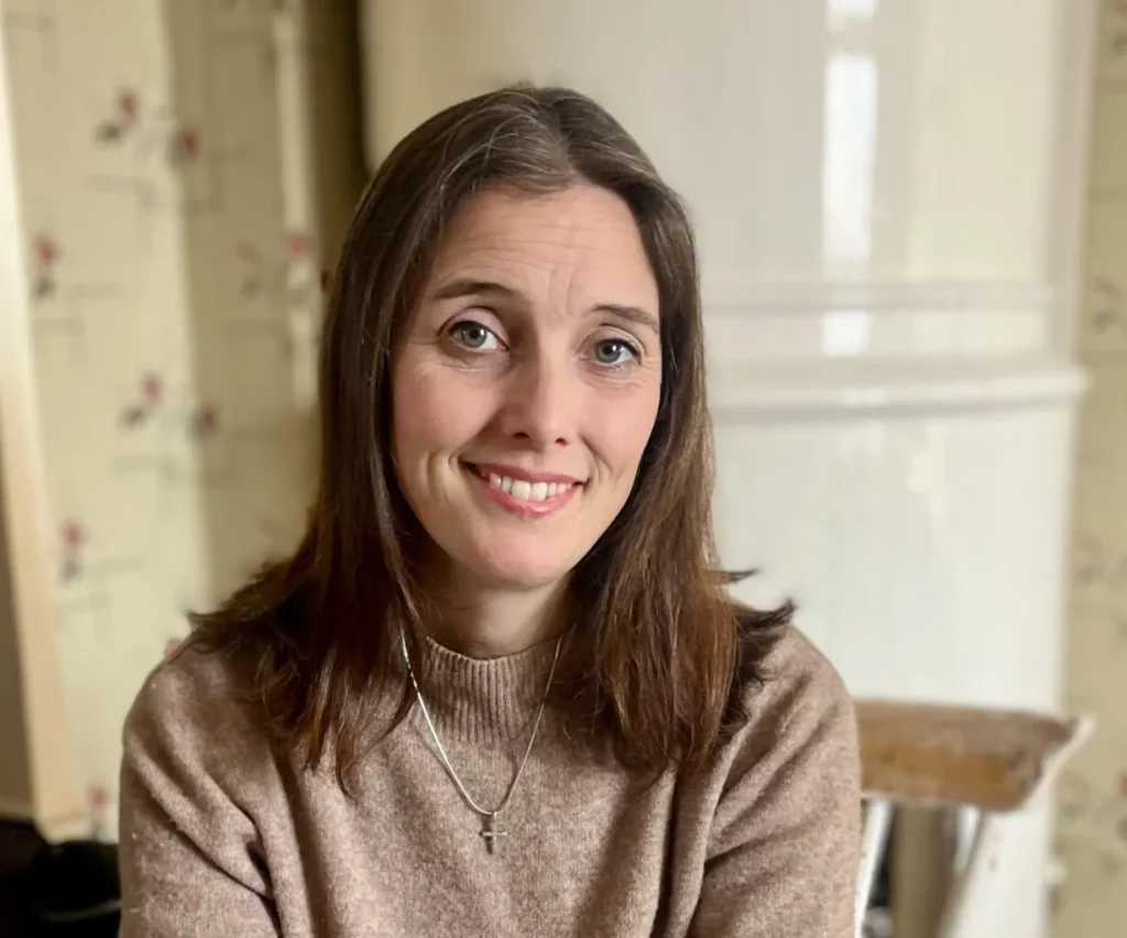 Kvinna med brunt hår och beige tröja ler inomhus, med blommig tapet och vit bakgrund.