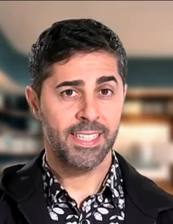 Man med kort mörkt hår och skägg, klädd i mönstrad skjorta och kavaj, ler inomhus.