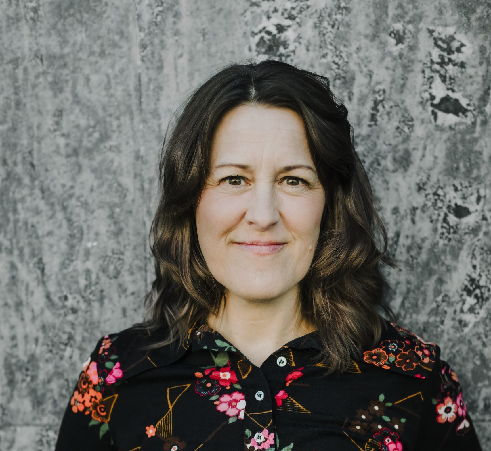 Kvinna med vågigt brunt hår, klädd i svartblommig skjorta, ler framför en grå marmorerad vägg.