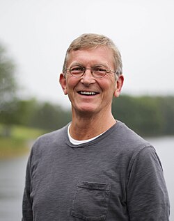 Leende äldre man med glasögon och grå skjorta som står utomhus nära vatten en molnig dag.