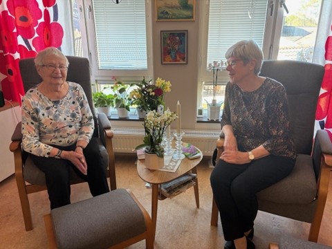 Två äldre kvinnor sitter och ler i ett mysigt vardagsrum med blommor och solljus som kommer in genom fönstren.