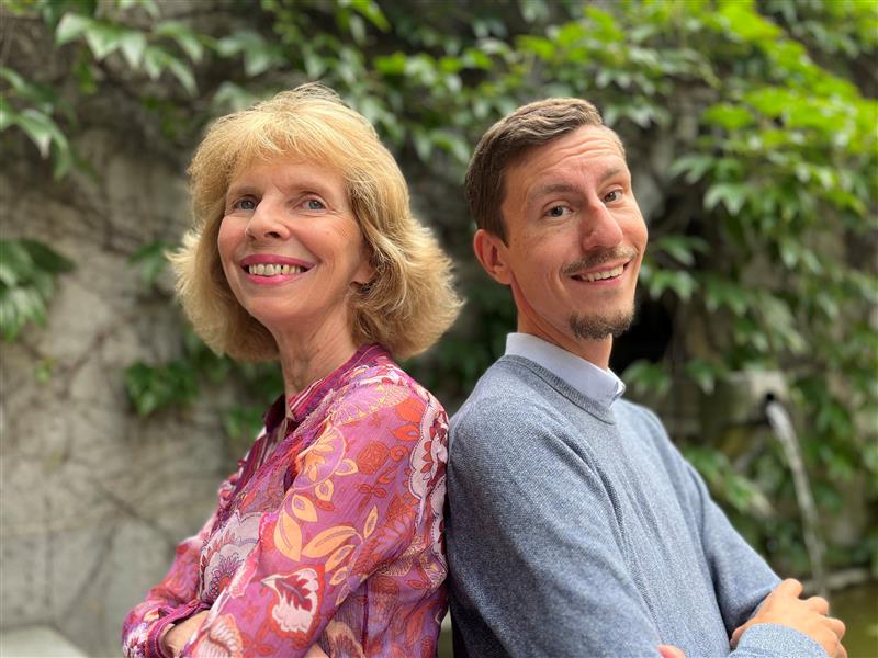 En leende kvinna och man står rygg mot rygg utomhus, med gröna blad i bakgrunden.