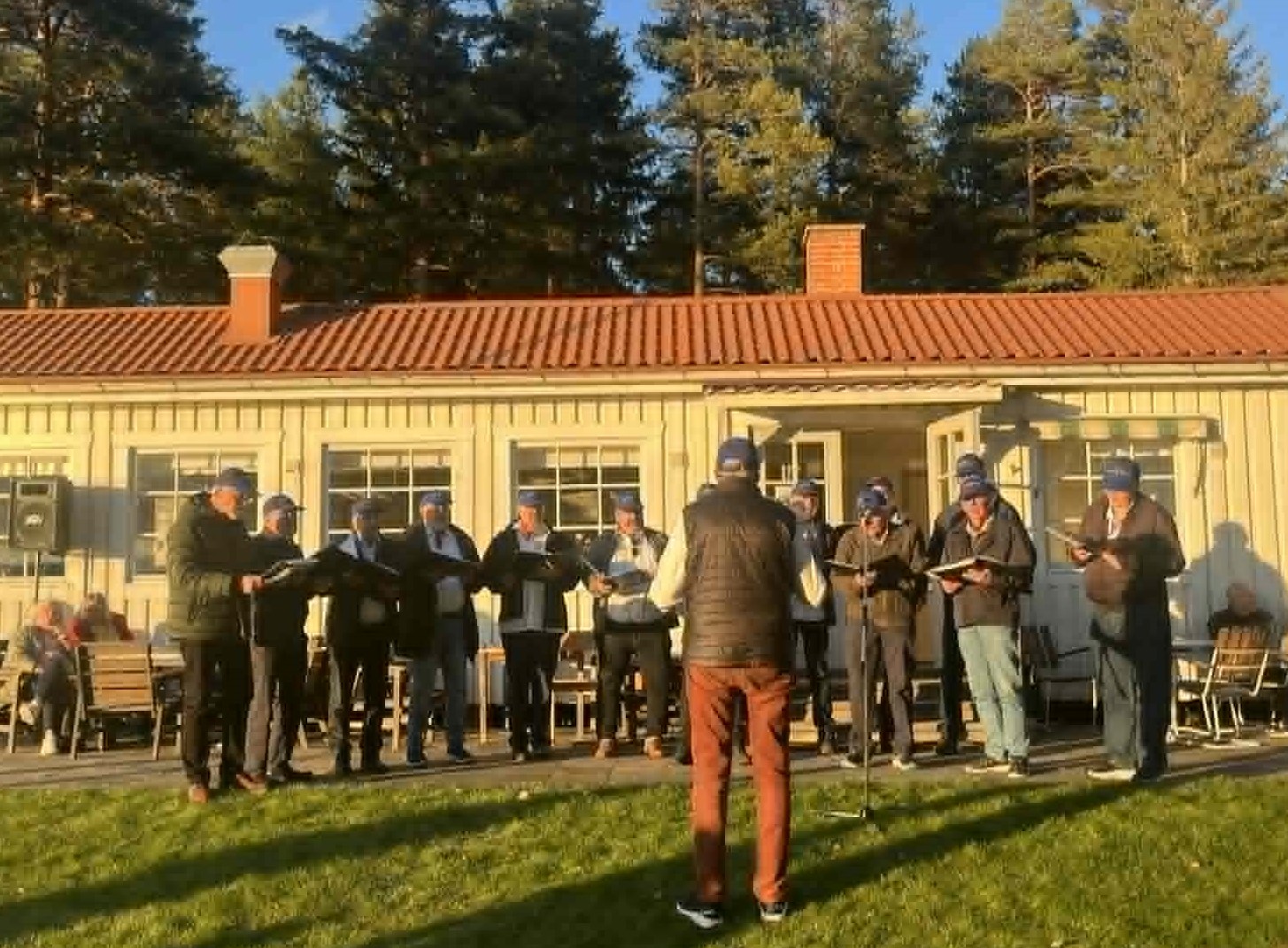 En grupp människor sjunger utomhus framför en träbyggnad med träd i bakgrunden.
