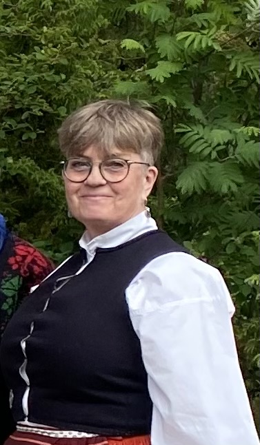 Leende person med glasögon, klädd i traditionella kläder, står utomhus framför grönt bladverk.