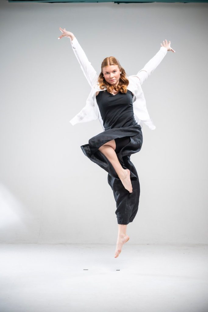 Rödhårig kvinna hoppar glatt i svart klänning och vit jacka mot en vit bakgrund.