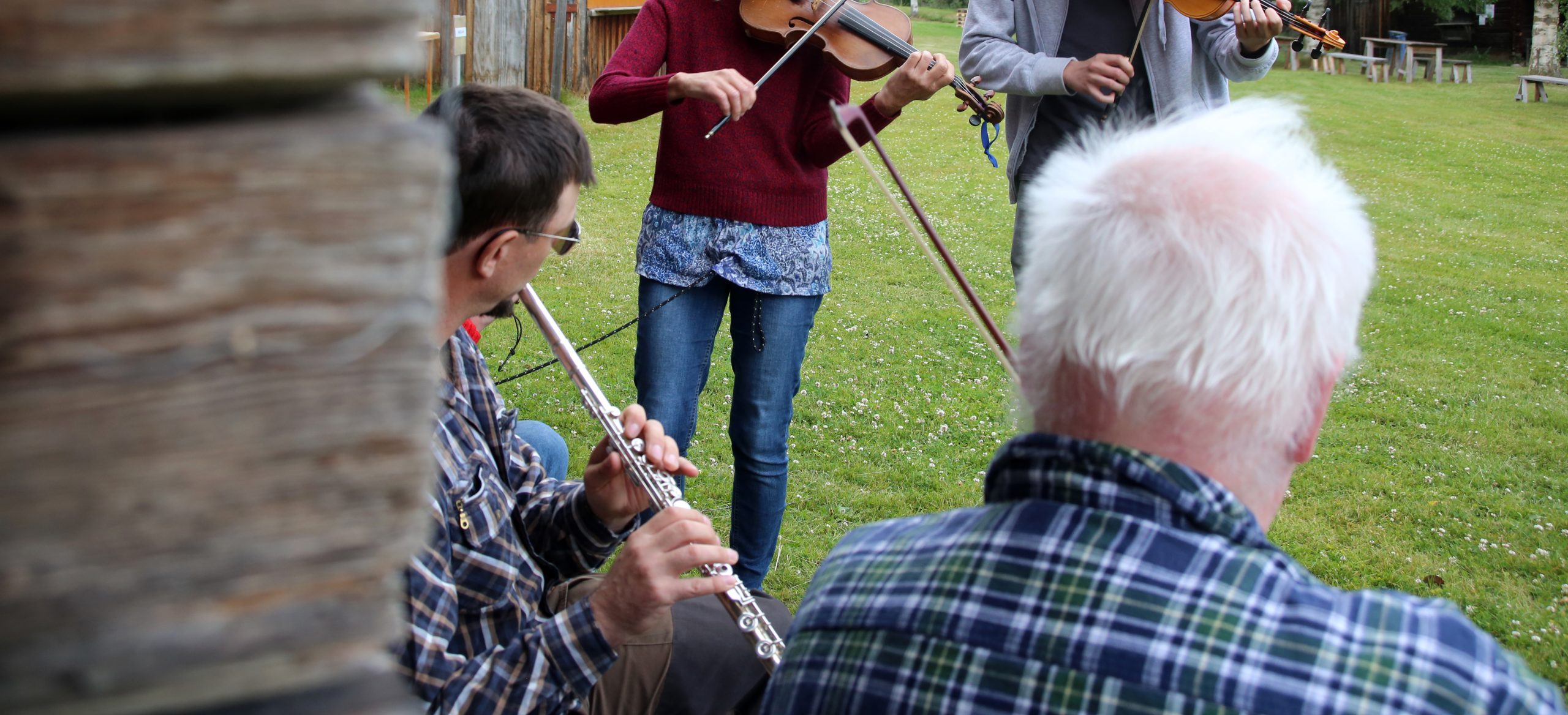Människor spelar flöjt och fioler utomhus på gräs, delvis bakom en träkonstruktion.
