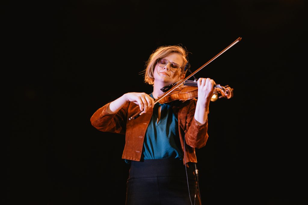 Person med glasögon spelar violin på scen, upplyst mot en mörk bakgrund.