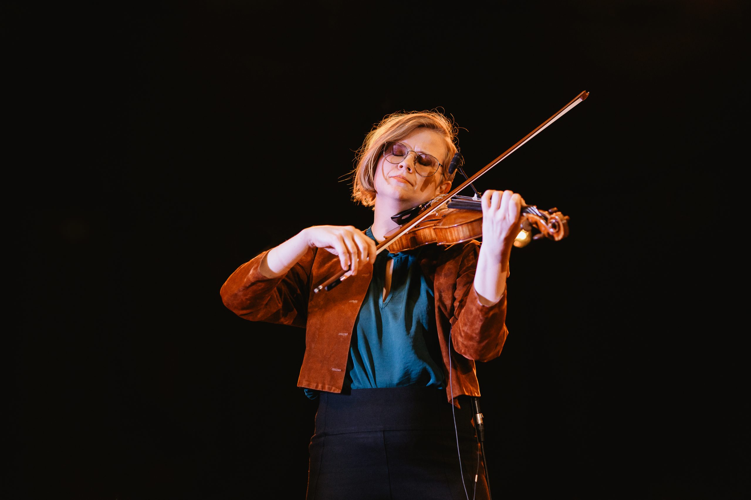 Person med glasögon spelar violin på scen, upplyst mot en mörk bakgrund.