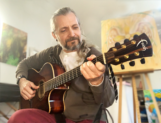 En man med långt hår spelar akustisk gitarr inomhus, med konst och målningar synliga i bakgrunden.
