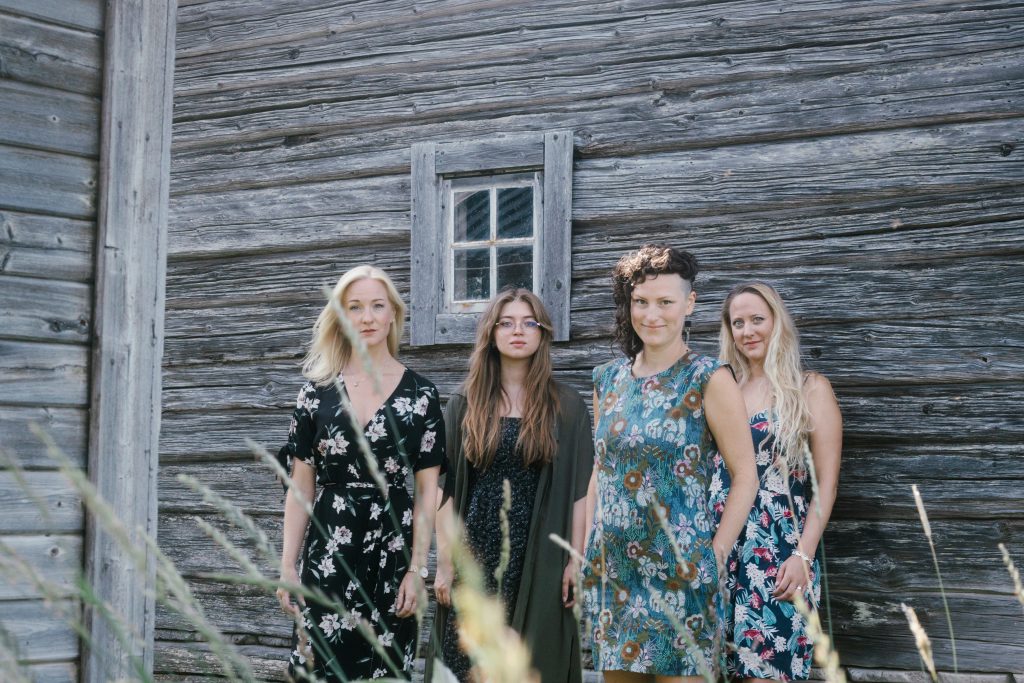 Fyra kvinnor i blommiga klänningar står framför en rustik trävägg med ett litet fönster.