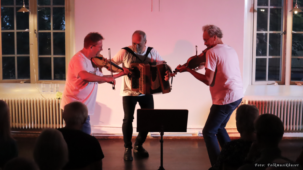 Tre musiker spelar violin och dragspel på scenen framför en publik, upplyst av varm inomhusbelysning.