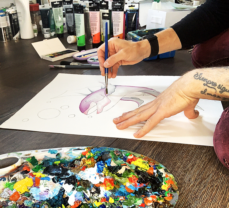 En person målar en delfin på papper, omgiven av färgtuber och en färgglad palett.