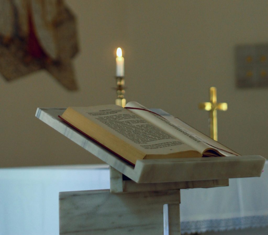 En öppen bok på ett stativ med ett tänt ljus och ett guldkors i en kyrkomiljö.