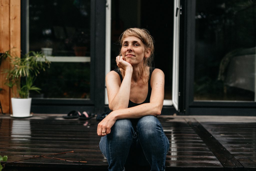 Kvinna sitter på en träveranda, vilar hakan på handen och ler eftertänksamt.