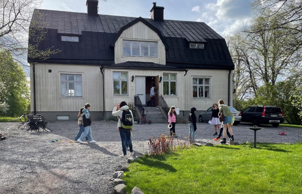 Barn och vuxna samlas utanför ett stort beigefärgat hus med svart tak en solig dag.