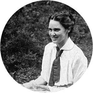 En mörkhårig kvinna i vit blus och slips sitter utomhus och ler.