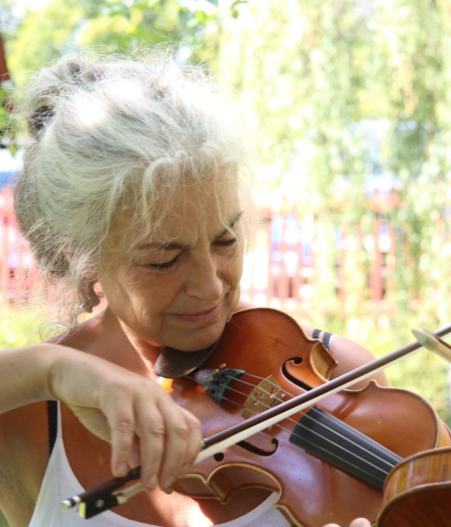 En äldre kvinna med grått hår spelar violin utomhus.