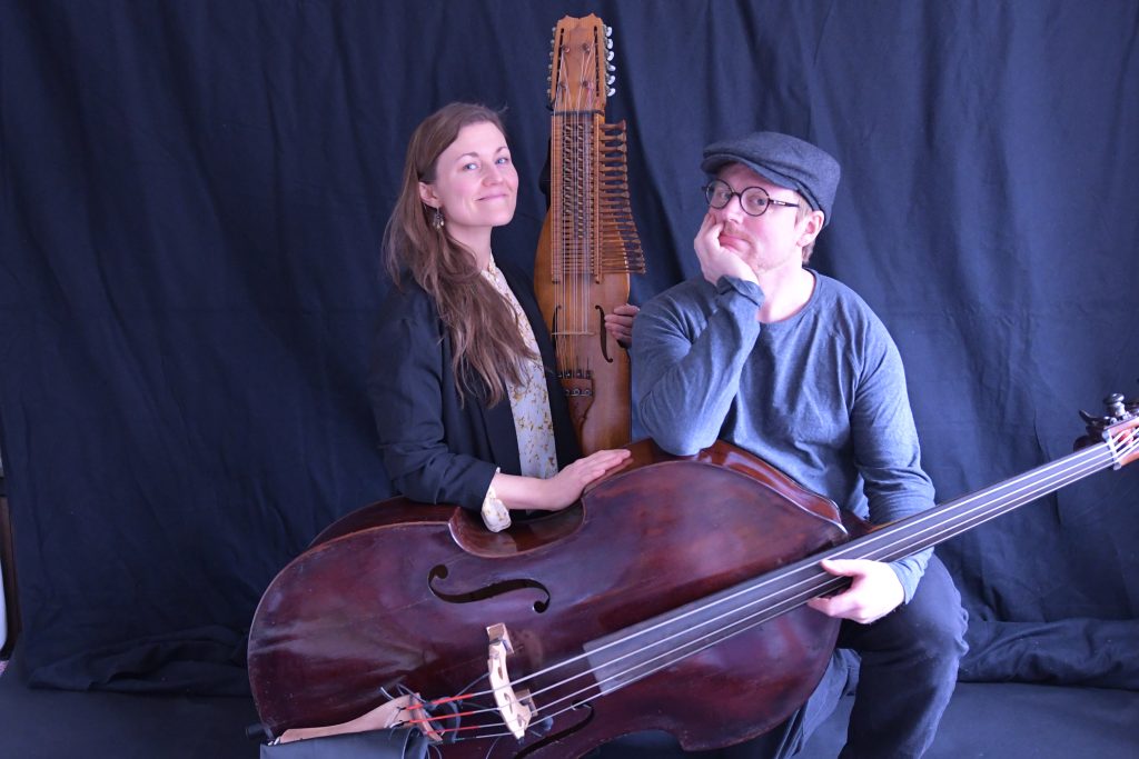 En kvinna och en man med nyckelharpa och kontrabas poserar framför en svart bakgrund.