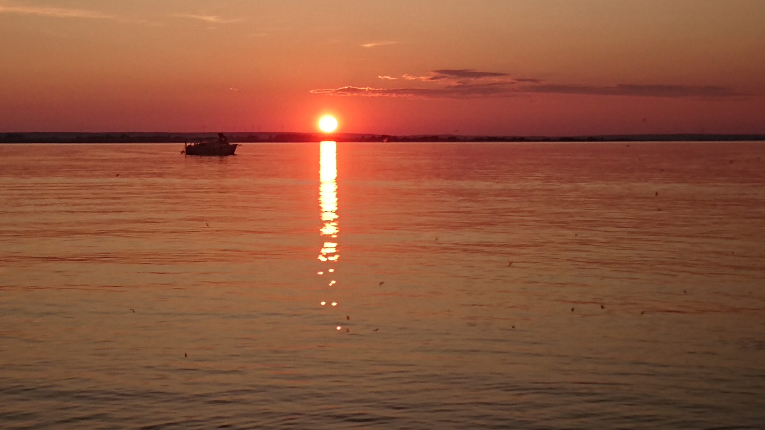 En båt på lugnt vatten i solnedgången, med solens reflektion som sträcker sig över den orangefärgade himlen och havet.