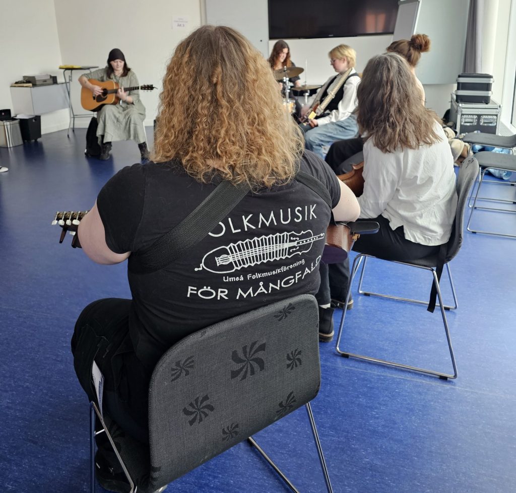 Människor sitter i en cirkel och spelar gitarr i ett ljust klassrum, en person har "Folkmusik" på sin tröja.