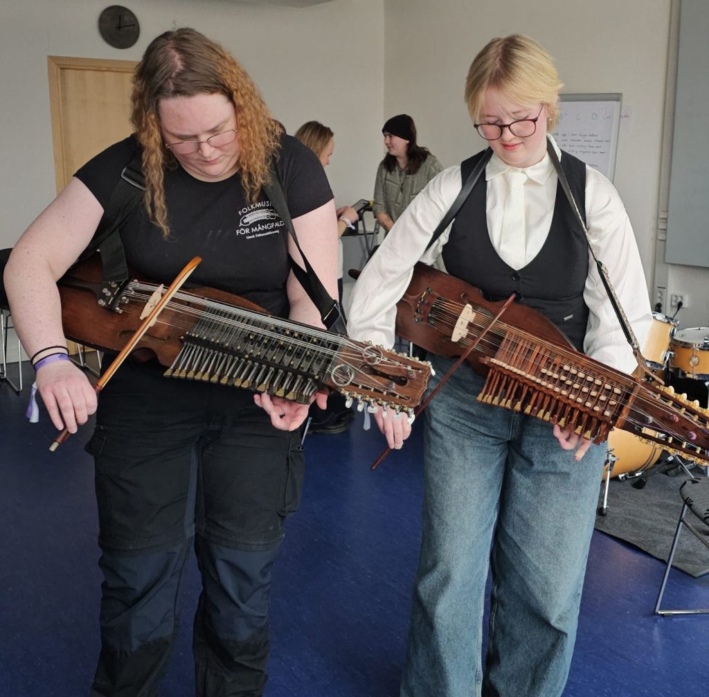Två personer spelar nyckelharpa i ett musikrum, med andra och trummor i bakgrunden.