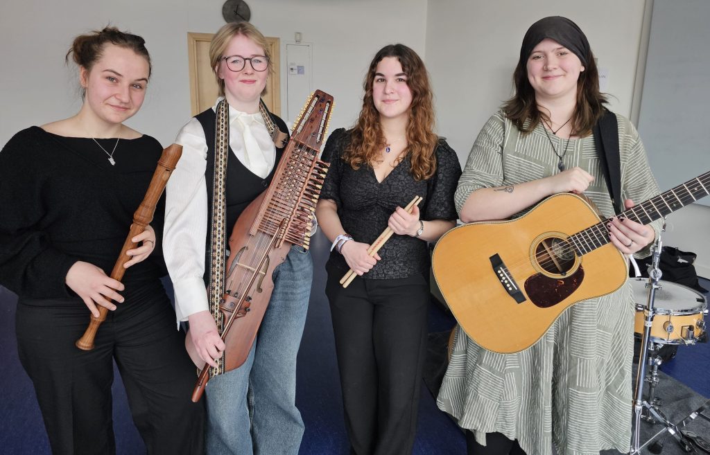 Fyra kvinnor står inomhus med musikinstrument: blockflöjt, nyckelharpa, trumpinnar och en akustisk gitarr.