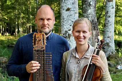 En man håller i en nyckelharpa och en kvinna håller i en fiol och poserar utomhus framför träd.