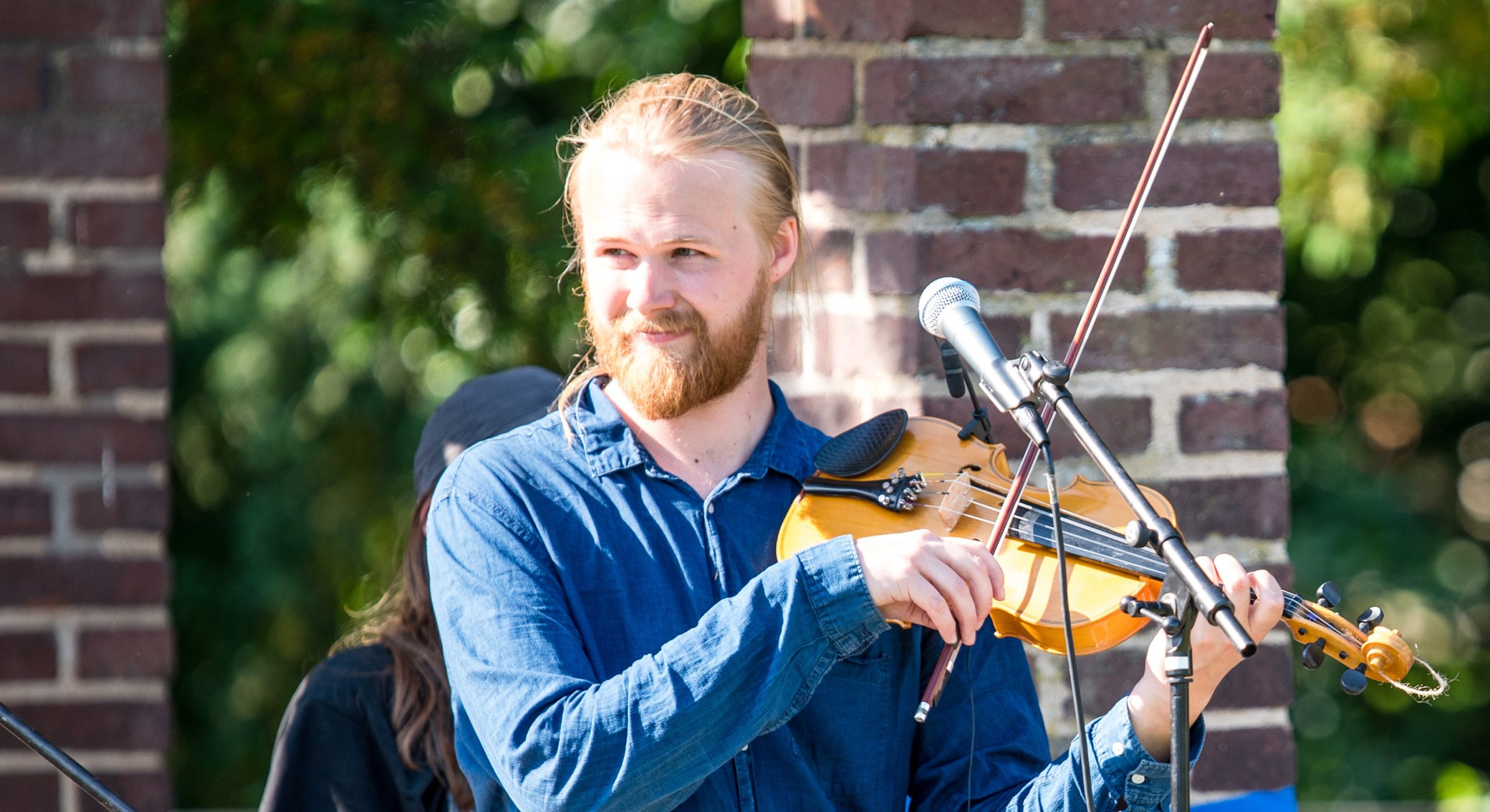 En man i blå skjorta spelar violin utomhus vid en murad pelare, med en mikrofon framför sig.