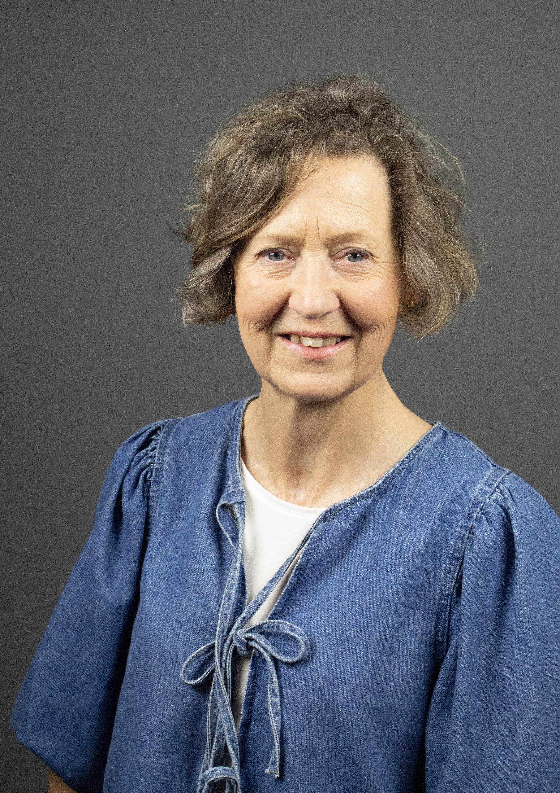 Leende äldre kvinna med kort, vågigt hår, klädd i blå jeansblus, poserar mot grå bakgrund.