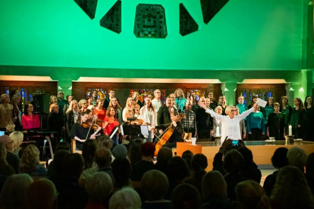 Kör och musiker uppträder på scenen i en kyrka med grönt ljus medan dirigenten gestikulerar till publiken.