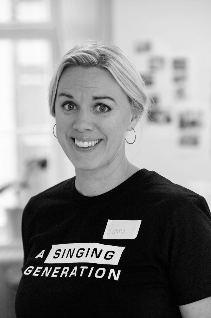 Leende kvinna med kort blont hår iförd en "A Singing Generation"-t-shirt och en namnskylt.