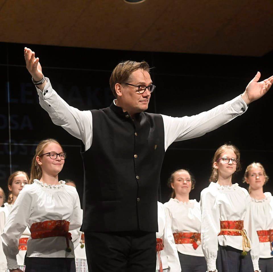 En man med armarna i vädret står framför flickor i traditionella kläder, som alla ler på scenen.
