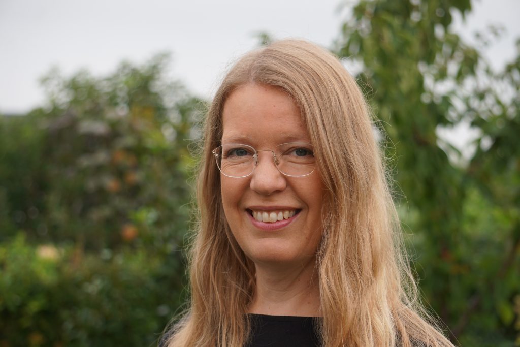 En kvinna med långt blont hår och glasögon ler utomhus med gröna träd i bakgrunden.
