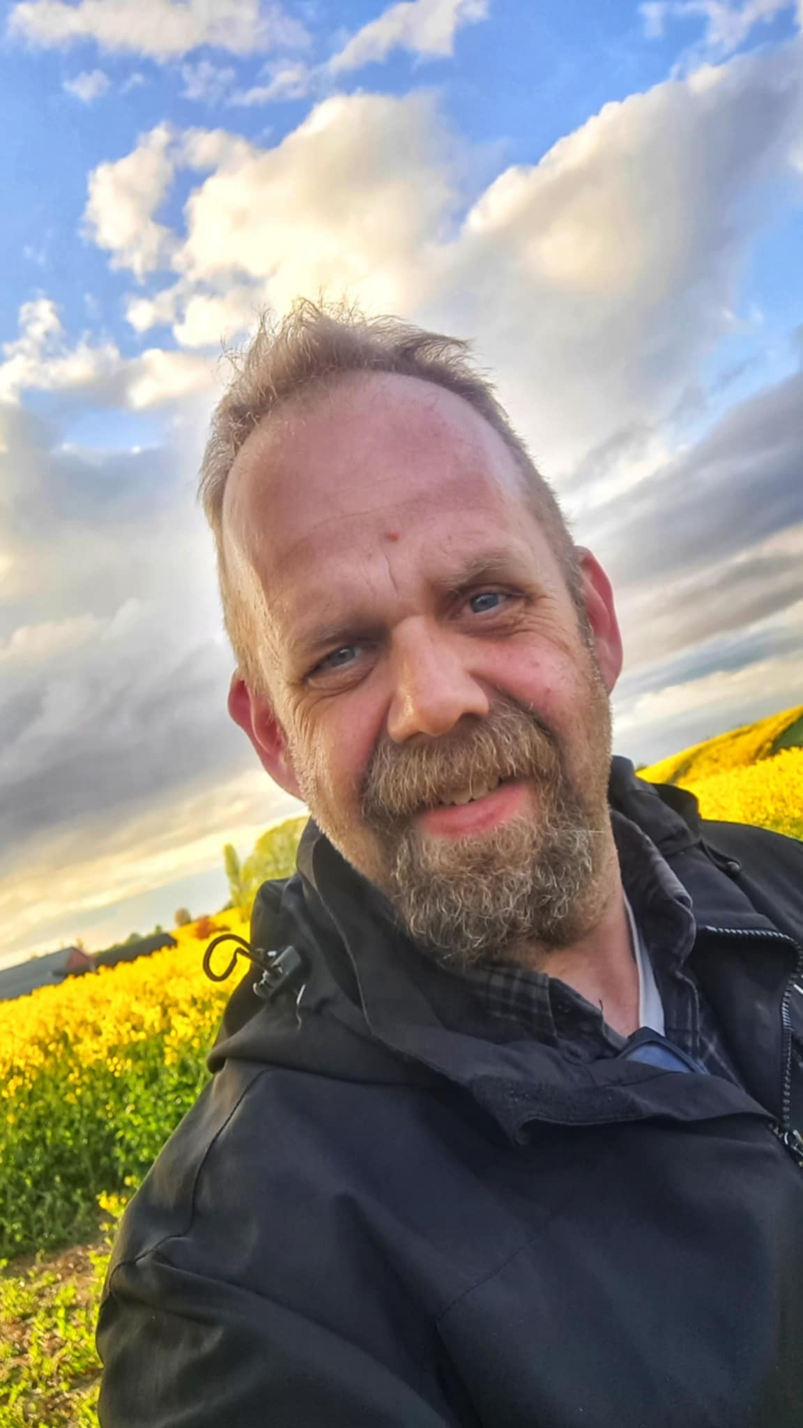 Leende man med skägg i svart kavaj står i ett gult blomsterfält under en molnig himmel.