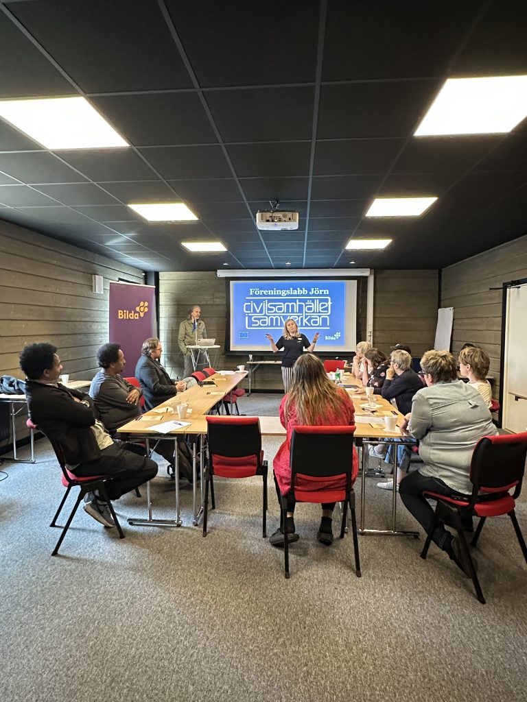 En grupp människor sitter runt bord i ett mötesrum och lyssnar på en presentatör via en skärm.