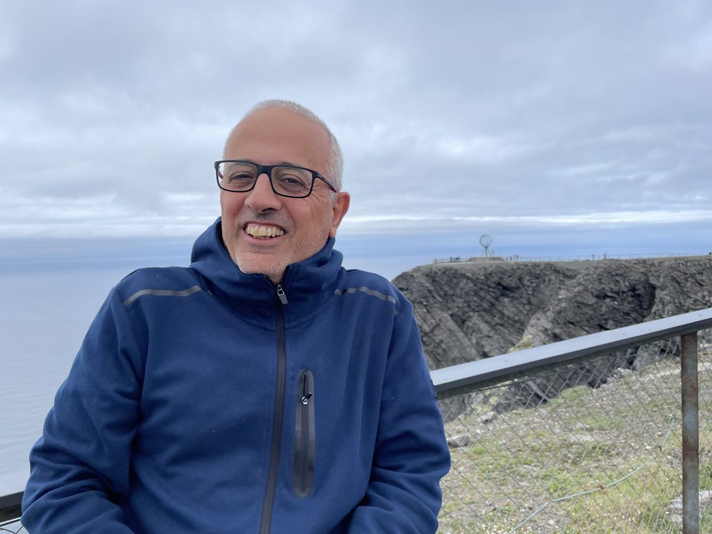 En leende man i blå jacka står vid en klippa med havet och en molnig himmel i bakgrunden.
