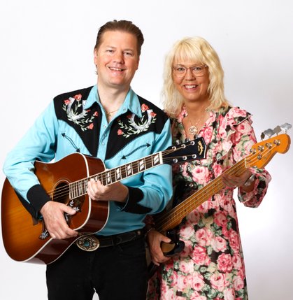 Två leende personer med instrument, den ena spelar akustisk gitarr, den andra håller i en basgitarr.