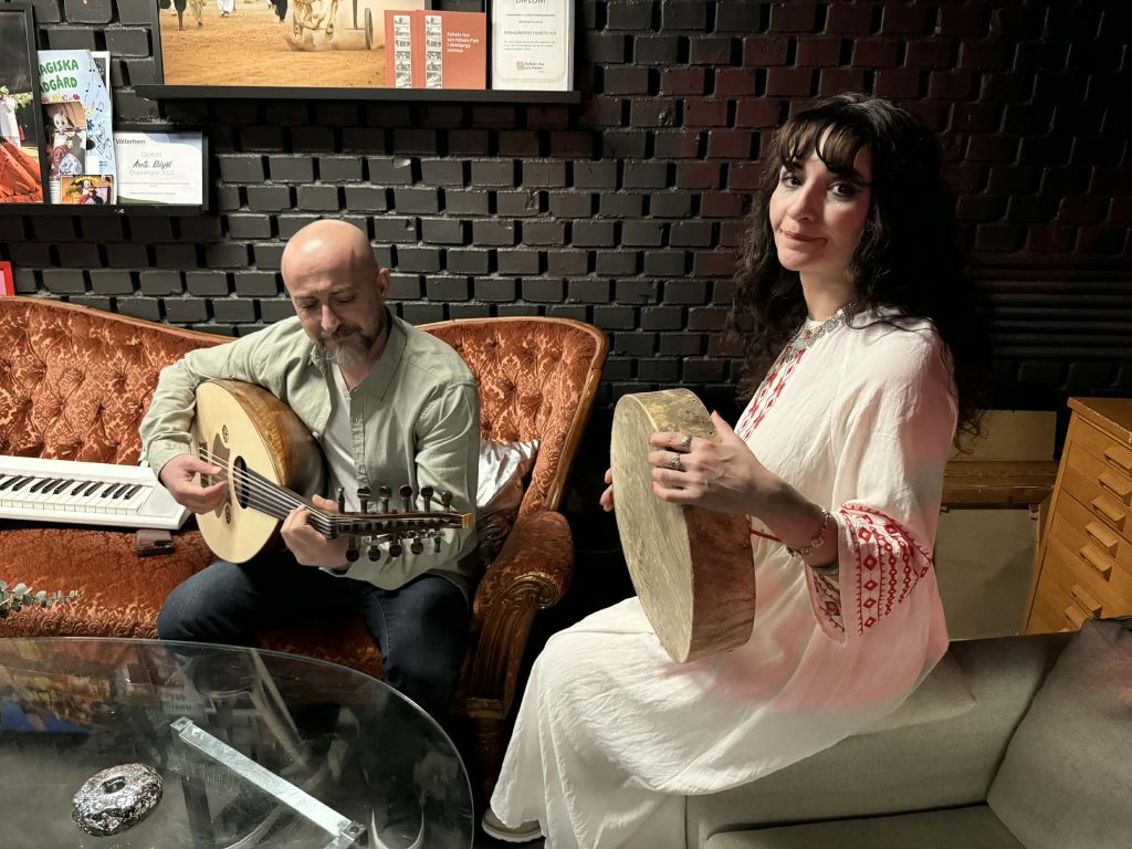 En man spelar oud och en kvinna spelar ramtrumma medan de sitter i en vintagesoffa i ett mysigt rum.